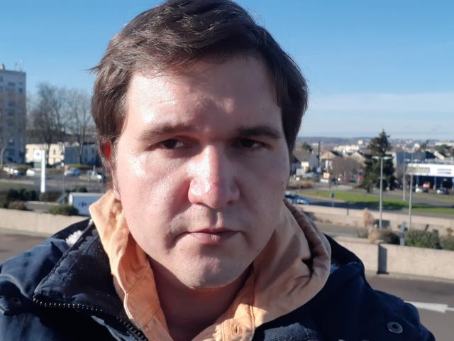 NectarineFad a un visage rond avec une peau pâle, des cheveux bruns et une expression sérieuse. Il porte une veste bleue avec une chemise de couleur pêche. Son nez est légèrement brillant..