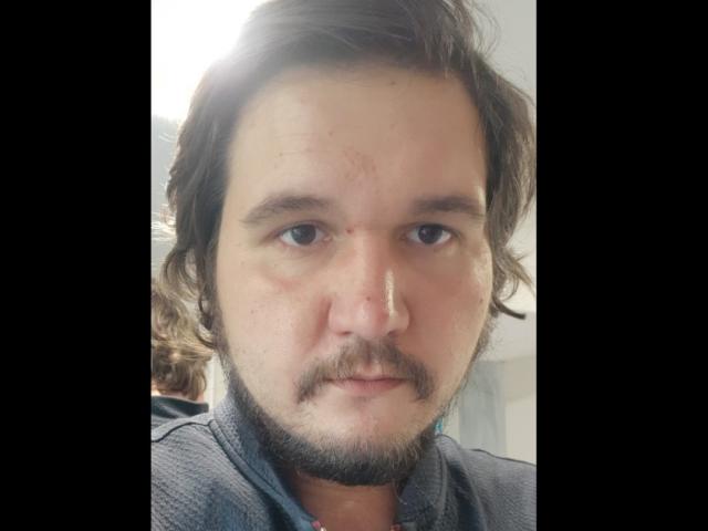 NectarineFad tem uma constituição média, cabelo castanho claro ondulado, uma barba curta e um pequeno corte acima da sobrancelha direita. Ele está vestindo uma camisa cinza..