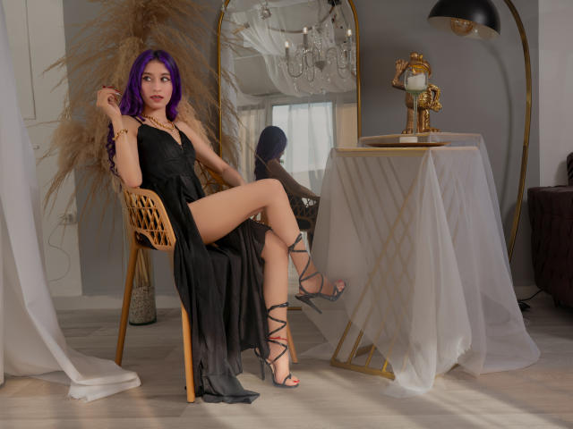 JuliaJobs with purple hair, black dress, and strappy heels, sitting on a wooden chair, showing her bare ass, next to a mirror and a gold lamp on a table.