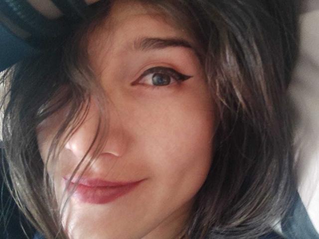 Le visage d'AliceMiz, avec ses cheveux brun foncé, légèrement en désordre, qui cachent un œil, son eye-liner noir et son sourire discret, révélant sa peau claire et ses lèvres rosées..
