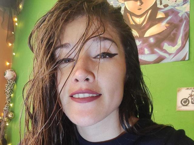 AliceMiz with wet brown hair, black eyeliner, pink lipstick, smiling, green wall, portrait of another person above, wearing black shirt, slight nose bump, soft light.