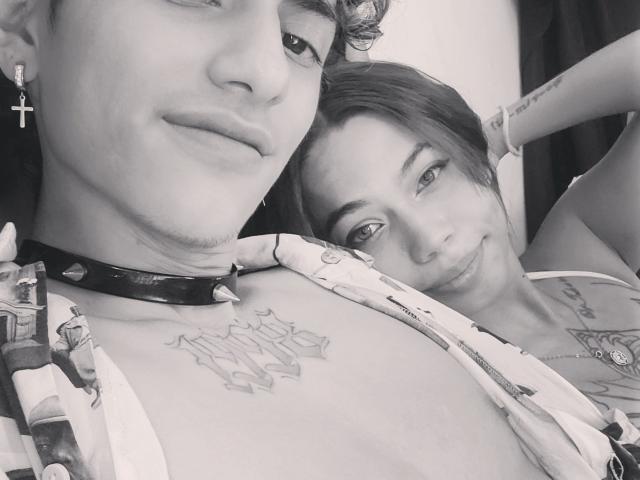 Selfie em preto e branco de AllucanTaste69 com tatuagens, usando um colar com espinhos e uma camisa estampada, apoiada em outra pessoa, AllucanTaste69, também com tatuagens e um bracelete. Ambas sorrindo..