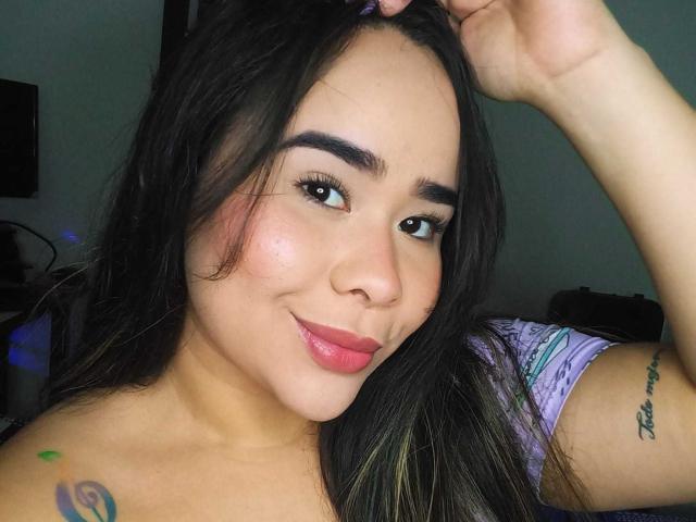 EveNailah, con cabello largo y negro, ojos marrones, labios rosados y un tatuaje en su brazo derecho, sonriendo y sosteniendo su cabello. Lleva una camisa morada..