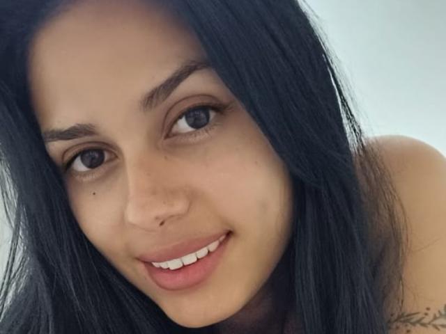AnaisHilton has dark, straight hair, brown eyes, and a small nose piercing. She's smiling with pink lips and a tiny beauty mark near her right eye.