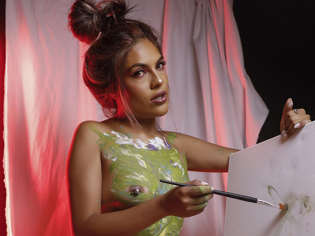 SunriseX with messy brown hair in a bun, painted green tits, holding a brush, painting white on a canvas, red light on her right, looking at the camera.