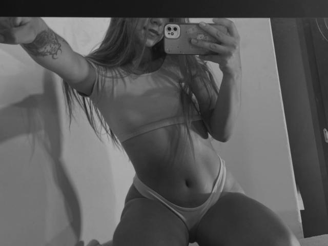 Black-and-white selfie of JuliaGarciax with long hair, wearing a crop top and panties, showing her flat stomach, small tits, and a tattoo on her left arm.