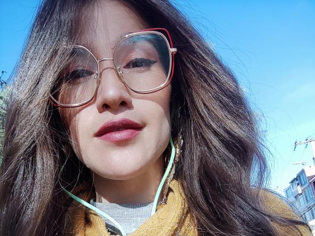 AliceMiz with wavy brown hair, red-framed glasses, and red lipstick, wearing a yellow hoodie, green earbuds, and a grey t-shirt, against a bright blue sky.