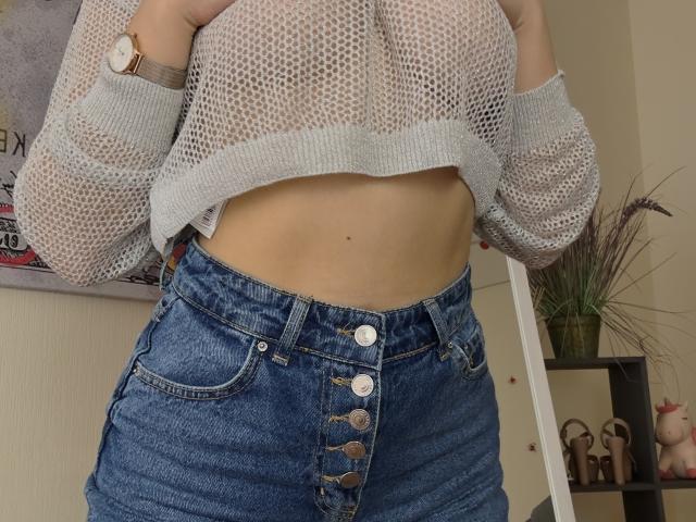 SugarrX wearing a sheer gray knit top, exposing her flat stomach and button-up high-waisted blue denim shorts with visible buttons. She has a gold watch on her wrist.