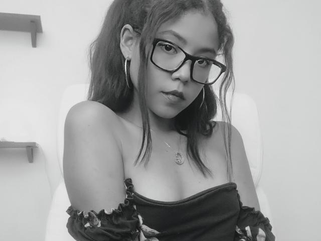 Zwart-wit foto van MaddieBlossom met een bril, oorringen en een top zonder bandjes, waarbij haar blote schouders en kleine borsten zichtbaar zijn, kijkend recht in de camera..