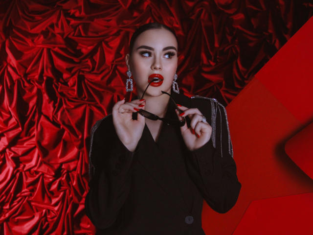 EricaWings with red lipstick, black dress, holding glasses in her mouth, red nails, earrings, and a serious expression, against red satin backdrop, looking to the side.