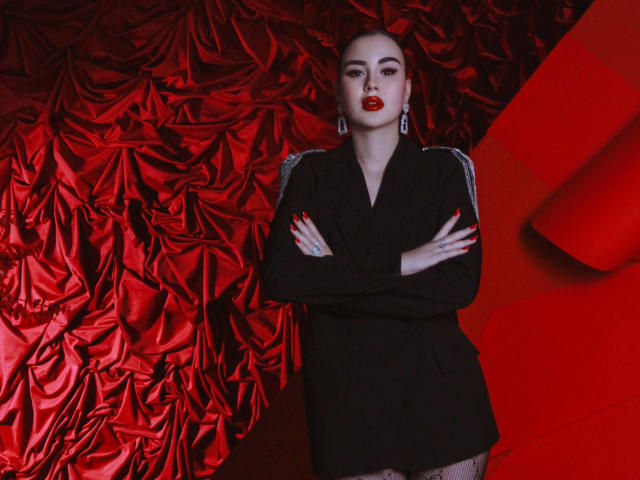 EricaWings stands with arms crossed, wearing a black dress, red lipstick, and earrings, against a red, textured, ruched background. Her hands have red nails and a ring.