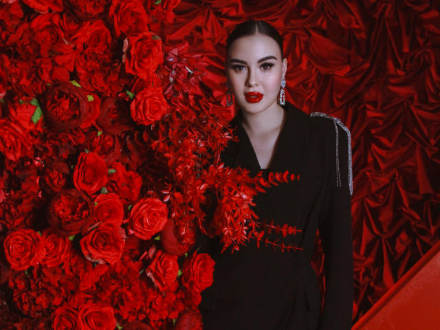  EricaWings with red lipstick, black dress, and earrings, standing against a wall of red roses, her dark hair slicked back, her ass partially covered by the flower arrangement.