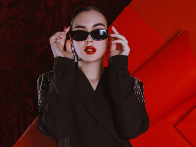 EricaWings with red lipstick, black sunglasses, and a black jacket with shiny embellishments, adjusting her glasses with both hands, looking confident and stylish.
