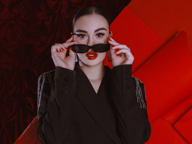 EricaWings with red lipstick, black sunglasses, and black jacket, holding glasses with red nails, wearing diamond earrings, looking at camera, red background, stylish, sexy, confident.