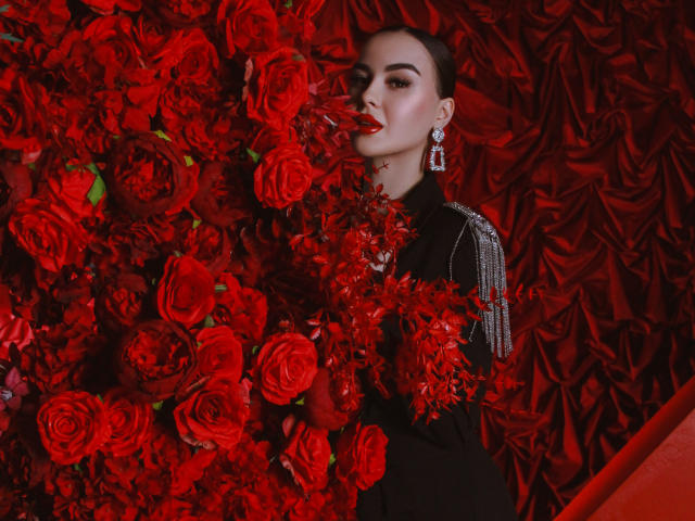 EricaWings with red roses in her mouth, black dress, and diamond earrings, holding more roses, looking sexy, red background, serious expression, her tits partially hidden by flowers.