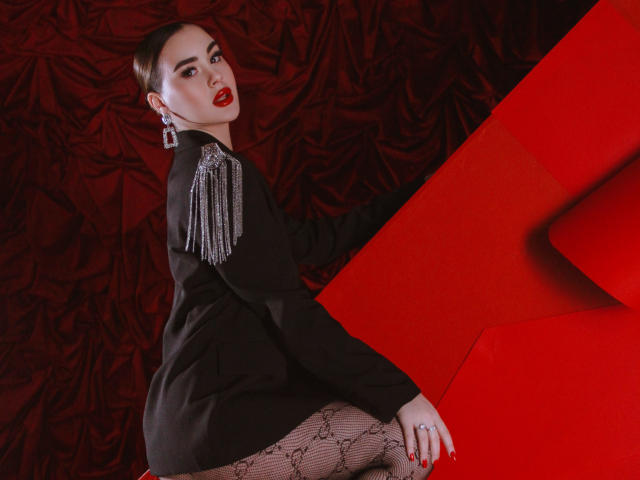 EricaWings with dark hair, red lipstick, black jacket with silver fringe, patterned tights, red geometric background, looking over shoulder, showing her ass, wearing large earrings.