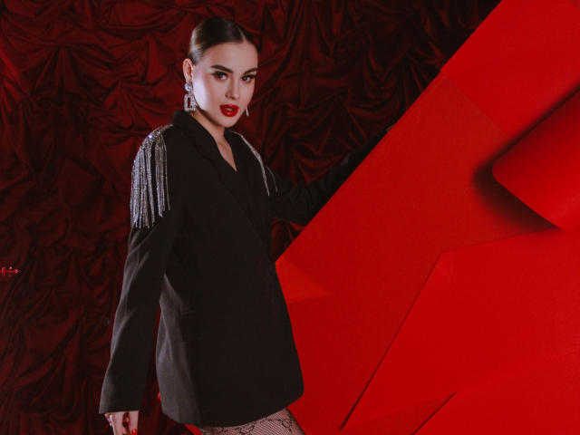 EricaWings with red lipstick, black jacket with silver epaulettes, and fishnet pants, standing against a red geometric pattern, looking directly at the camera.