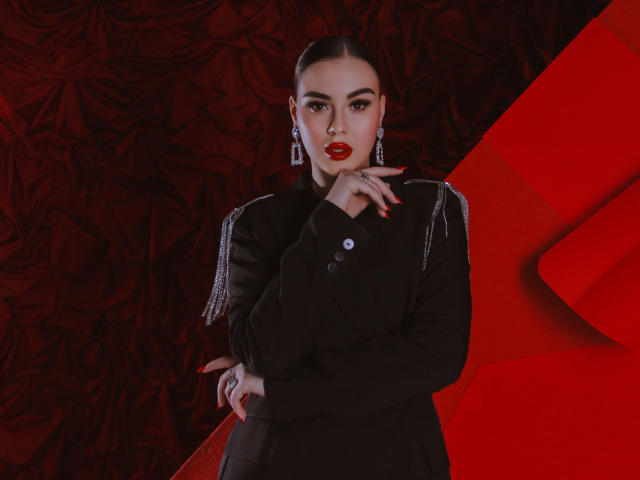 EricaWings has red lipstick, dark eyes, and is wearing a black button-up with silver fringes, touching her chin with red nails, and wearing dangling earrings.