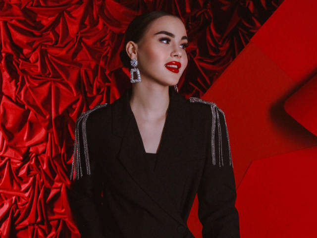 EricaWings with red lipstick, black blazer, and diamond earrings, standing against a red, textured background, looking to the side with a confident, sexy expression.