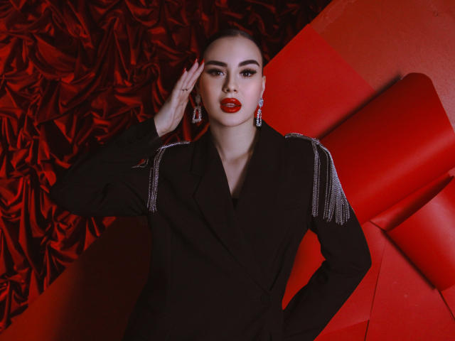 EricaWings has red lipstick, black jacket with silver fringes, and red earrings, saluting with right hand, standing against red and black background, looking fierce and sexy.