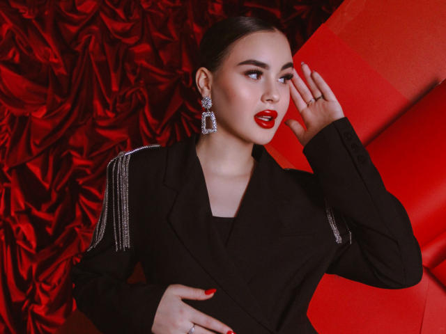 EricaWings with red lipstick, black blazer, and sparkly earrings, hand raised to face, red background, and red velvet texture, looking to the side, nails painted red.