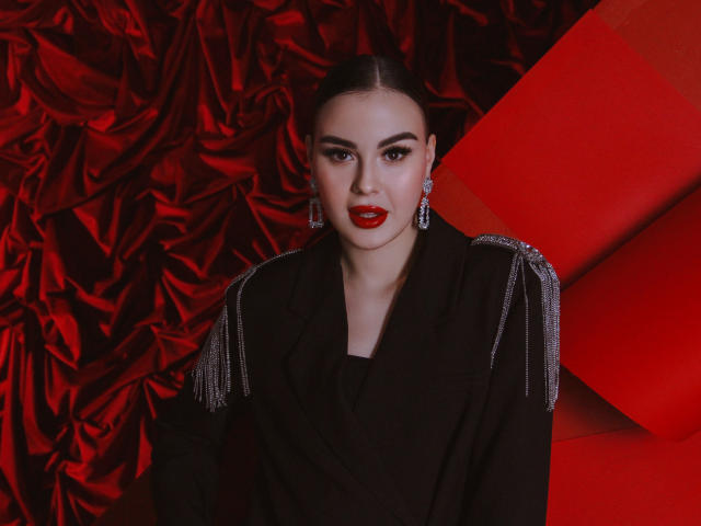 EricaWings with red lipstick, black jacket, and silver earrings, looking at camera with confident smile, dark hair in sleek bun, red background, shiny textures, stylish and elegant.
