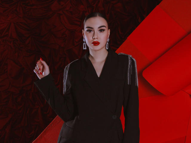 EricaWings with red lipstick, black jacket, and silver earrings, raising her manicured hand. She's got a serious face, and her tits are slightly visible under the jacket.