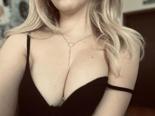 SugarrX with blonde wavy hair, black deep cleavage dress, and a delicate necklace, showing her big, full tits. Soft, natural light.