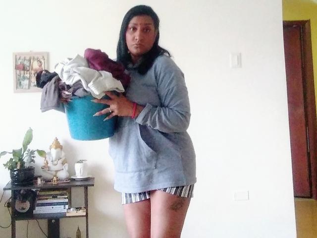 Nahirr, a dark-skinned woman with medium-length black hair, wearing a gray hoodie, holds a blue laundry basket with clothes, showing her striped underwear and tattoo on her right thigh.