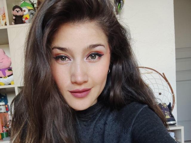 AliceMiz a de longs cheveux bruns, des yeux verts avec du crayon noir, un fard à paupières rose et du rouge à lèvres rose. Elle porte un pull noir à col roulé, elle sourit légèrement, avec un fond blanc..