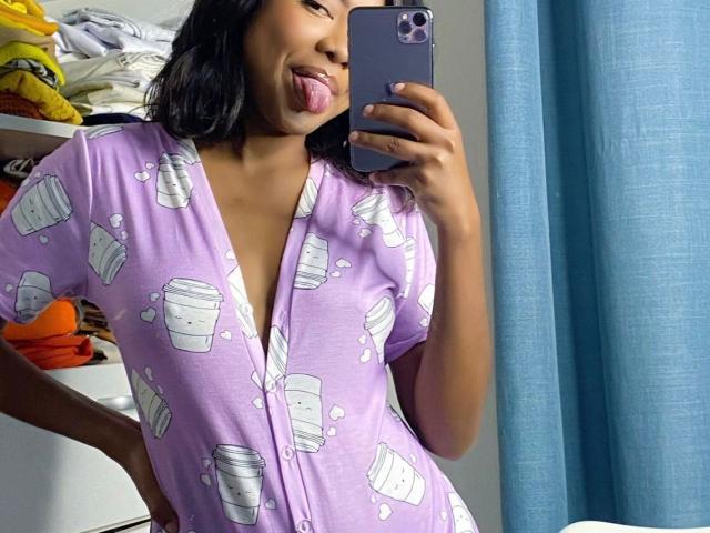 BabeHottie69 with dark hair, sticking out tongue, wearing purple pajama top with white cupcake prints, taking mirror selfie with purple phone, showing deep V-neck, cute and playful.