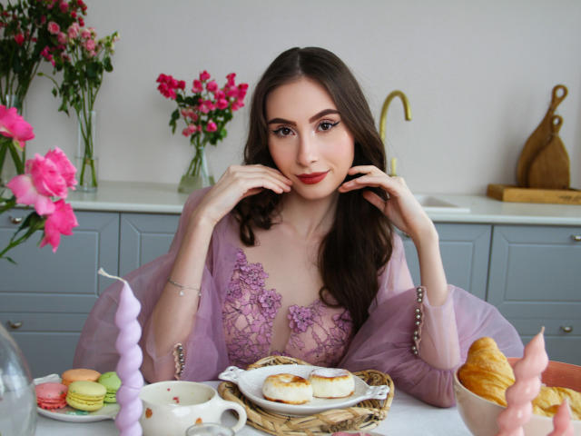 SaraQuincy, avec ses longs cheveux bruns, sa robe en dentelle violette et son rouge à lèvres rouge, assise à une table avec des pâtisseries, des macarons et un décor de gâteau rose, a l'air mignonne et séduisante..
