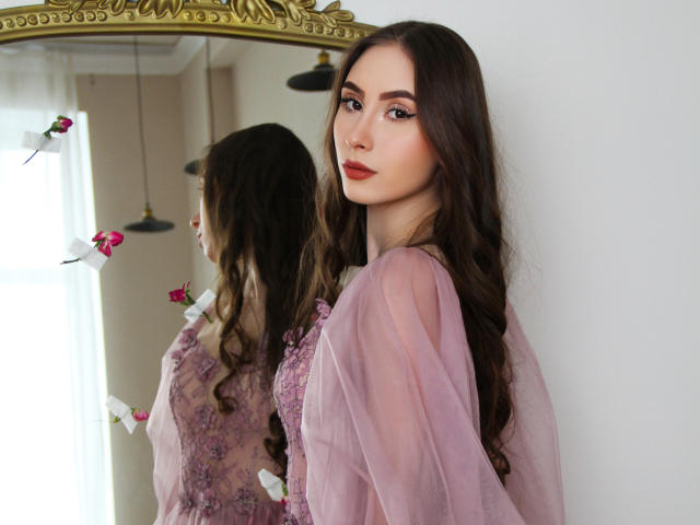 Sara Quincy, com longos cabelos castanhos, blusa rosa transparente com detalhes em renda, olhando para o espelho, o reflexo mostra seu perfil, batom vermelho, luz suave, rosas cor-de-rosa ao fundo, expressão suave..