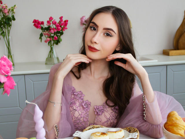 SaraQuincy, avec ses longs cheveux bruns, sa robe en dentelle violette et son rouge à lèvres rose, se tient la main au menton, assise à une table avec des croissants et un ballon rose. Ses seins sont petits..