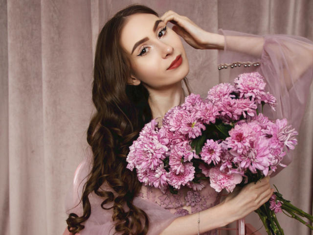SaraQuincy, com longos cachos castanhos, segurando crisântemos rosa, vestindo uma blusa rosa transparente, tocando a testa, batom vermelho, sorriso suave, fundo rosa delicado, elegante, sexy..