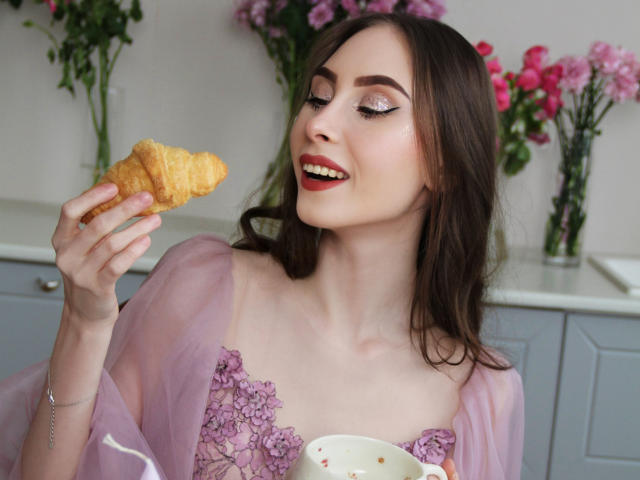 SaraQuincy, met volle borsten, houdt een gouden croissant vast, draagt een doorschijnende roze badjas en rode lippenstift, en glimlacht met gesloten ogen, terwijl ze een witte beker met een roze bloem vasthoudt..