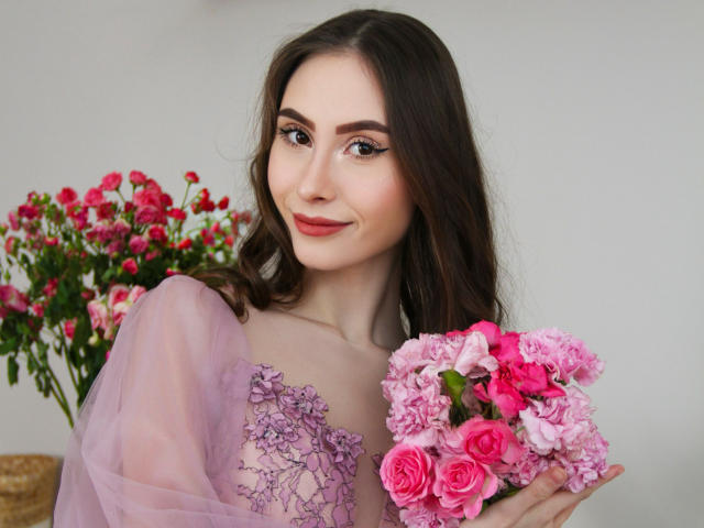 SaraQuincy con fiori rosa, che tiene un bouquet, indossa un abito viola trasparente con pizzo, sorridente, capelli castani ondulati, pelle chiara, rose rosa, garofani rosa, trucco leggero, carina..