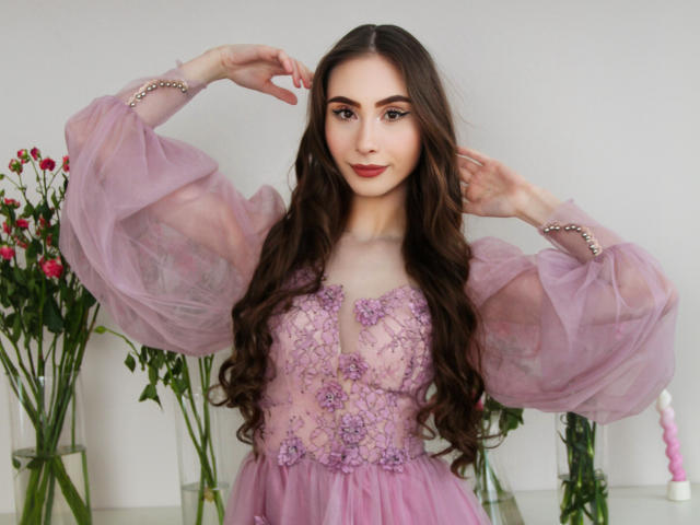 Sara Quincy, com cabelos castanhos longos, vestido de renda rosa, mangas bufantes, bordados florais, segurando o cabelo para cima, sorrindo, em pé em frente a flores, rosa e branco..