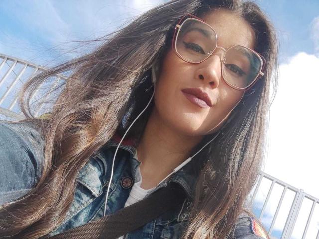AliceMiz with long brown hair, wearing a denim jacket, oversized glasses, and white earbuds, has a cute face and slightly parted pink lips, with a brown strap visible.