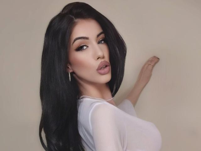 RavenRoseBerry, com cabelos pretos longos, lábios cheios e uma blusa branca, exibindo seus seios grandes contra uma parede lisa, olhando por cima do ombro com uma expressão sensual..