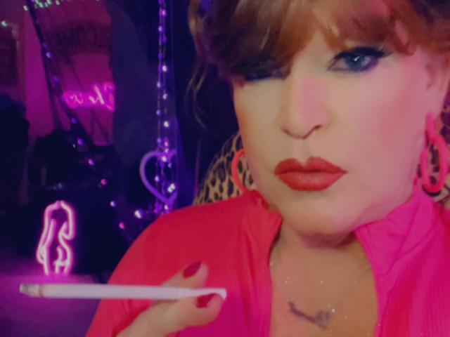 FemmeCheri with red lipstick, blue eye makeup, and pink jacket, holding a cigarette, wearing pink hoop earrings, and a necklace with a small cross.