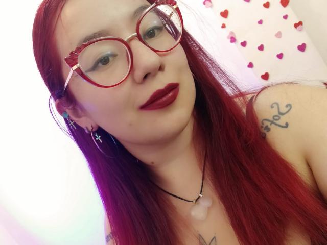 HollyManziniBBW with red hair, red glasses, red lipstick, tattoo on shoulder, necklace with white gem, looking at camera, wearing hoop earrings, showing upper chest and face.