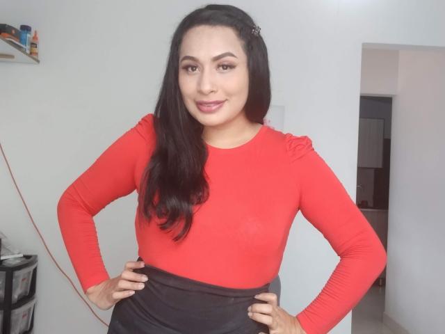 AlicTwenty, con lunghi capelli neri, indossa una maglia a maniche lunghe rossa e pantaloni neri, mani sui fianchi, sorridente, con un sedere piccolo, seno di media grandezza, pelle chiara, trucco leggero..