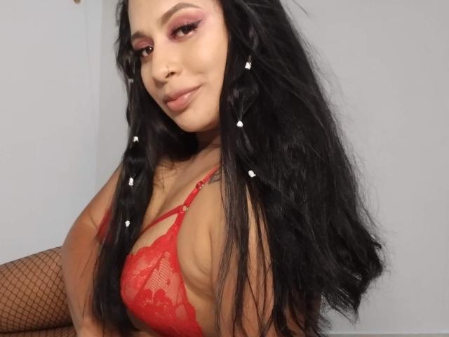 AlicTwenty with long black hair, wearing red lace bra and fishnet stockings, has pink eyeshadow, small nose ring, and a slight smile, showing her tits and a tattoo.