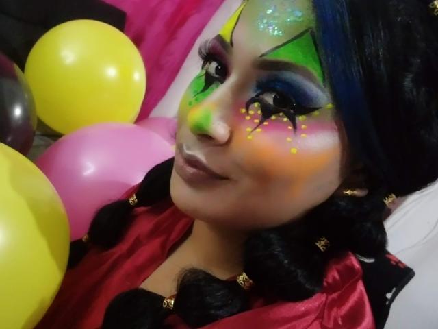 AlicTwenty with colorful face makeup, green, yellow, and purple eyeshadow, yellow and pink balloons, black braids, red satin shirt, and glossy lips, smiling at the camera.