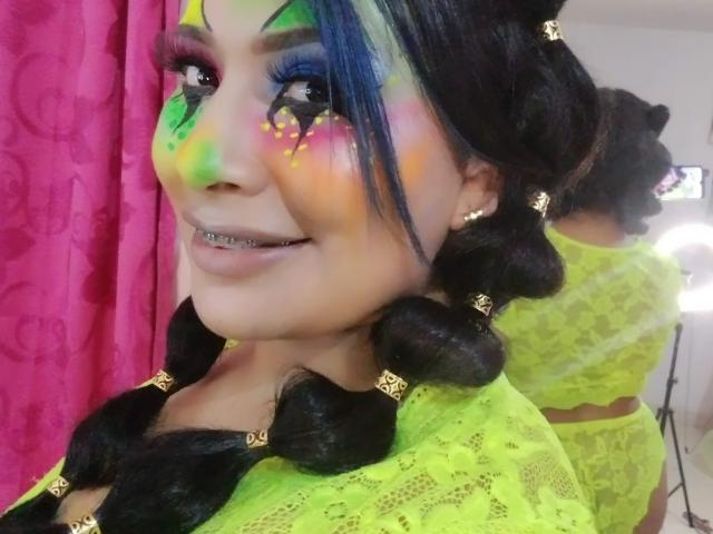 AlicTwenty con il viso dipinto con colori vivaci, una maglia di pizzo verde neon, trecce nere, fermagli dorati, che sorride mostrando denti metallici. Trucco vivace, tenda rosa, riflesso nello specchio..