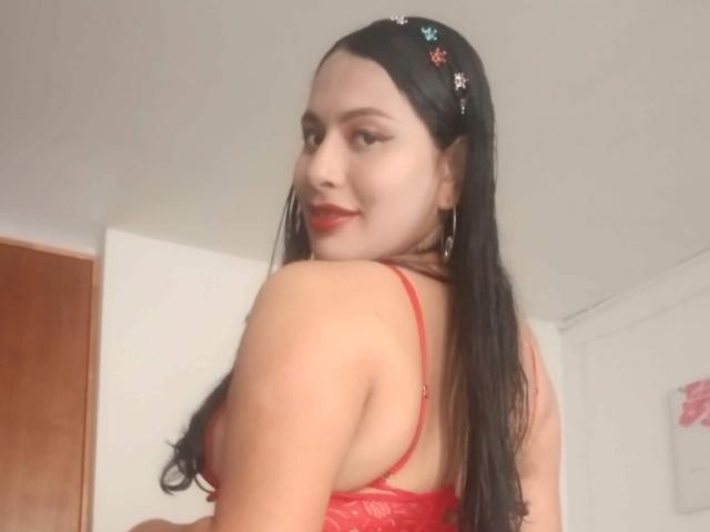 AlicTwenty with long black hair, red lace bra, and red lipstick, looking over her shoulder, smiling, wearing a floral headband, and small hoop earrings, in a bright room.