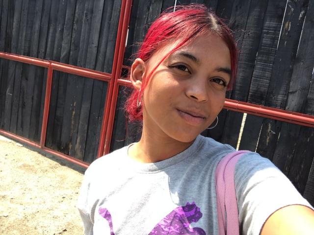 HilaryCook tiene el pelo rojo, piel de color marrón medio y pechos pequeños. Lleva una camisa gris con flores moradas, una correa rosa y está sonriendo en un selfie tomado con luz solar..