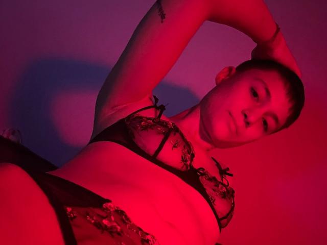 NaughtyBushyJessy in red-lit lingerie, arms above head, showing small tits in black lace bra, short brown hair, tattoo on arm, looking down with slightly parted lips.