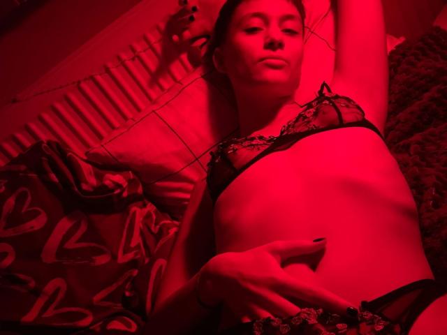 NaughtyBushyJessy deitada na cama, com iluminação vermelha, vestindo um sutiã e calcinha de renda preta, braços acima da cabeça, mão na barriga, com uma aparência sedutora, com os seios e a vulva ligeiramente visíveis..